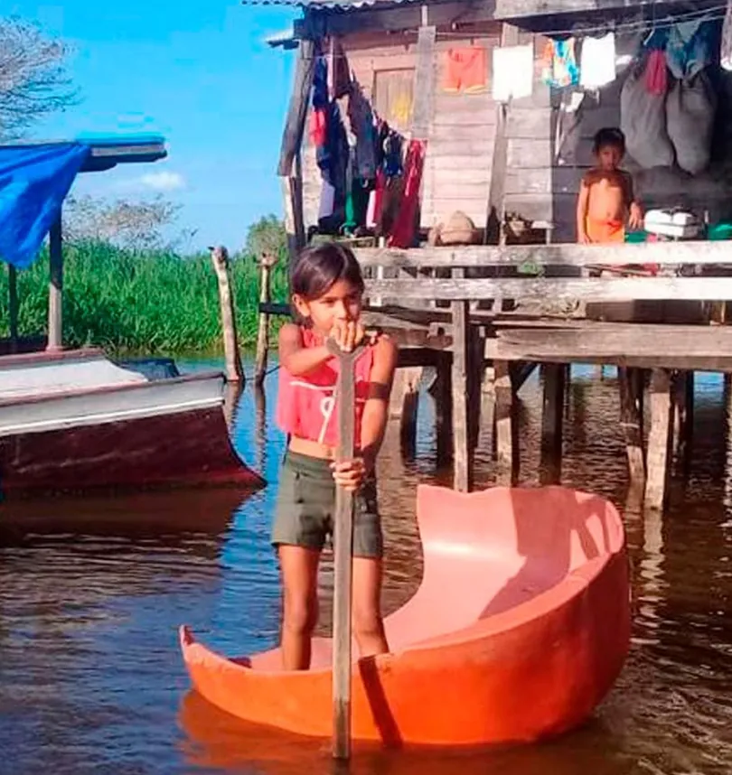 Menina remando uma pequena canoa próxima a uma casa ribeirinha